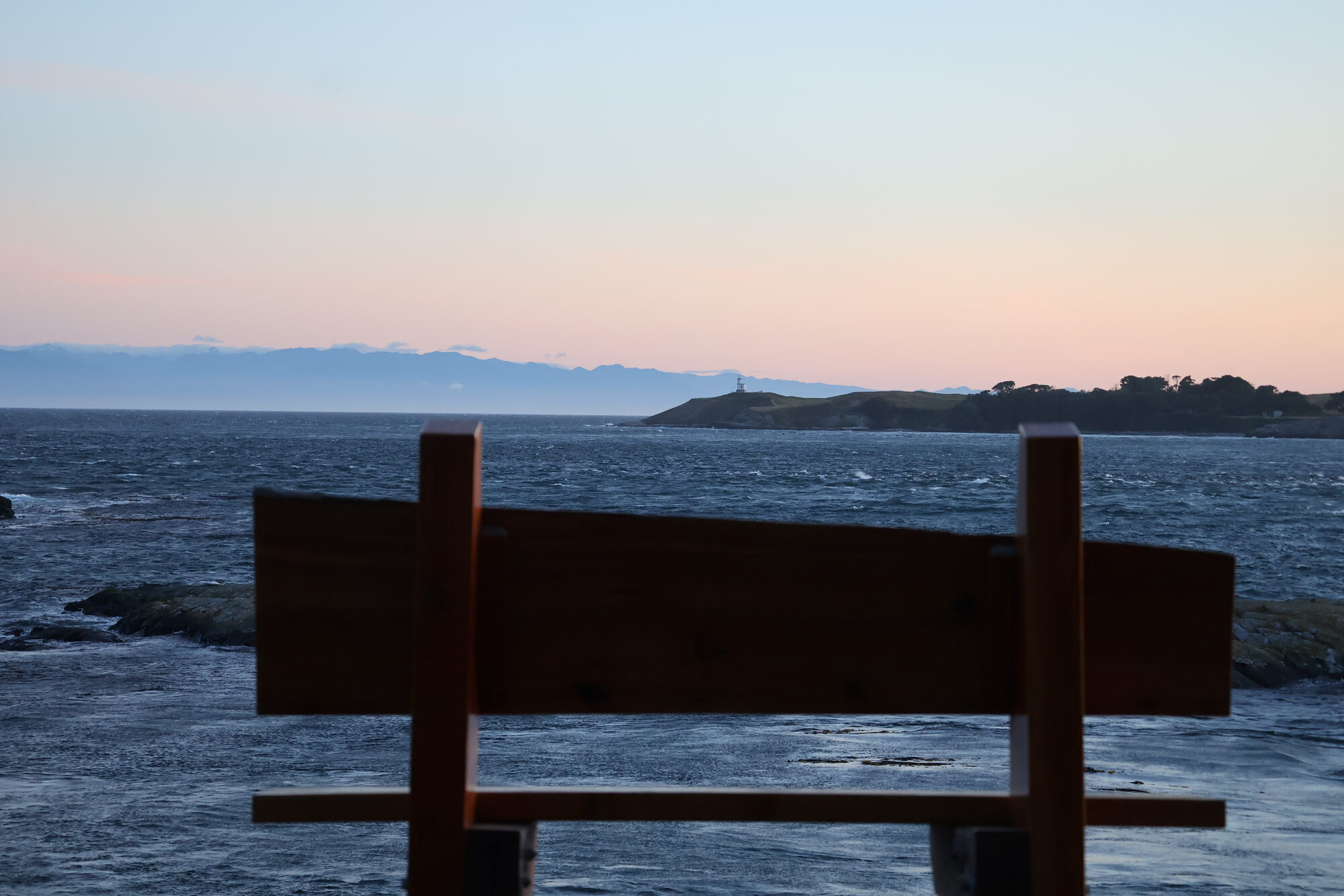 An empty wooden bench facing the sea at dusk, lighthouse in the distance — print